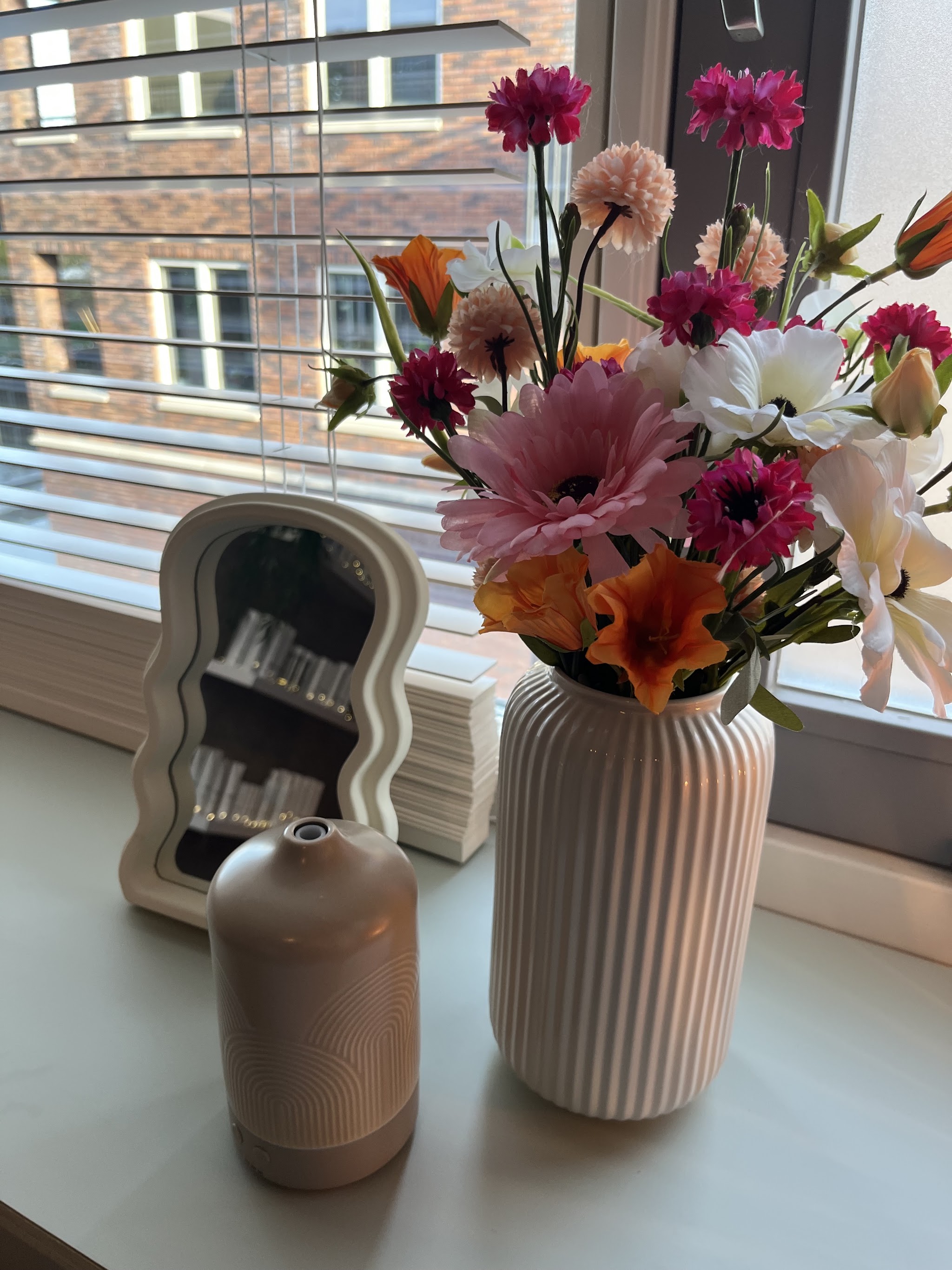 Bloemen in een geribbelde vaas, een golvend spiegeltje en een diffuser op de vensterbank van de salon